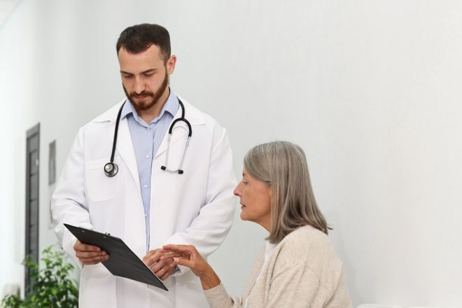 A doctor reviews a clipboard with an older woman in a clinic hallway.