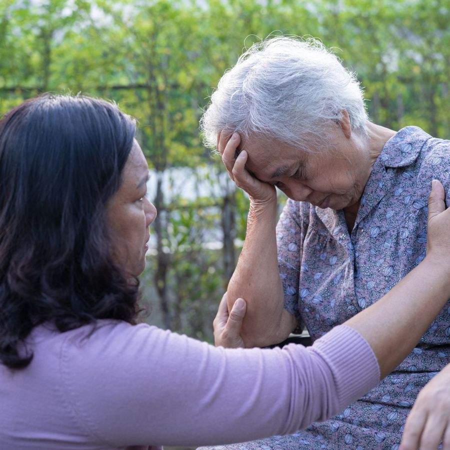 A woman kneels beside her mother in a garden, holding her arm and comforting her.