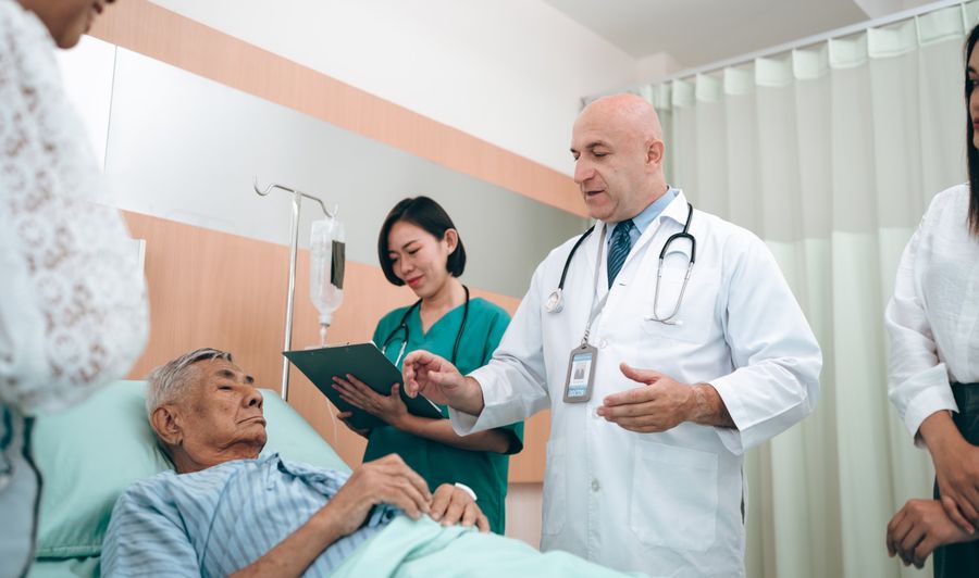 A doctor and nurse talk with an older man in a hospital bed while his family listens.
