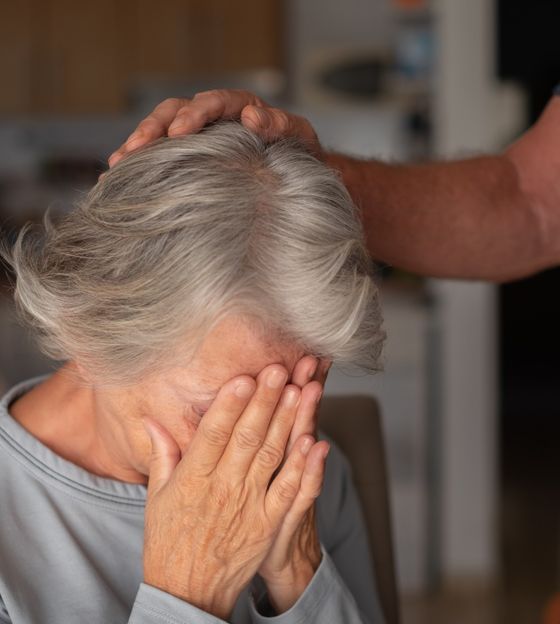 A family member rests a reassuring hand on the head of an older woman who covers her face.