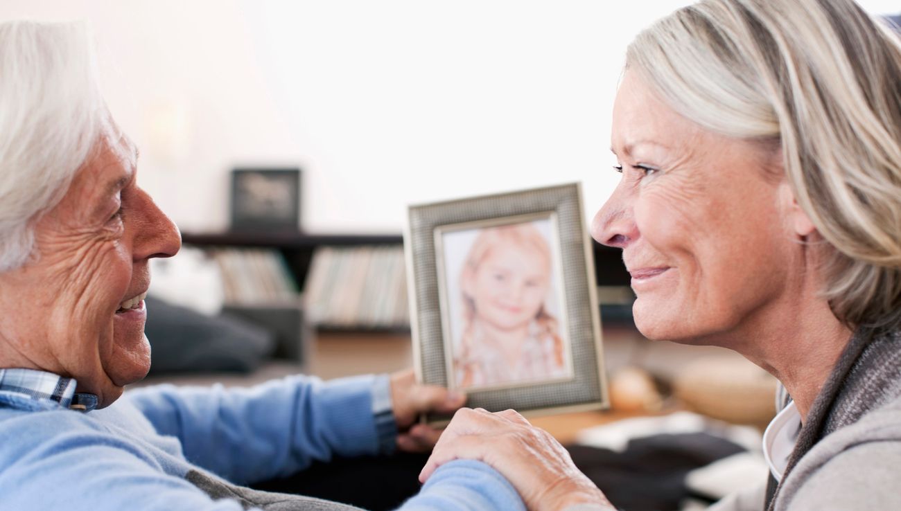 An older man and his daughter smile at each other while holding a framed photo of a child.