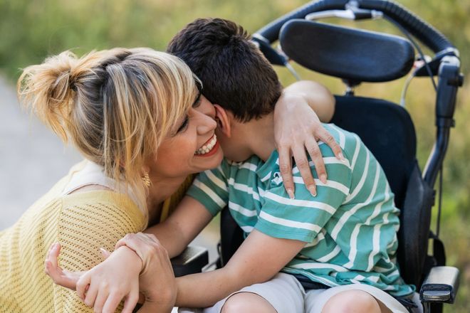 A smiling mother hugs her son, who sits in a wheelchair outdoors.