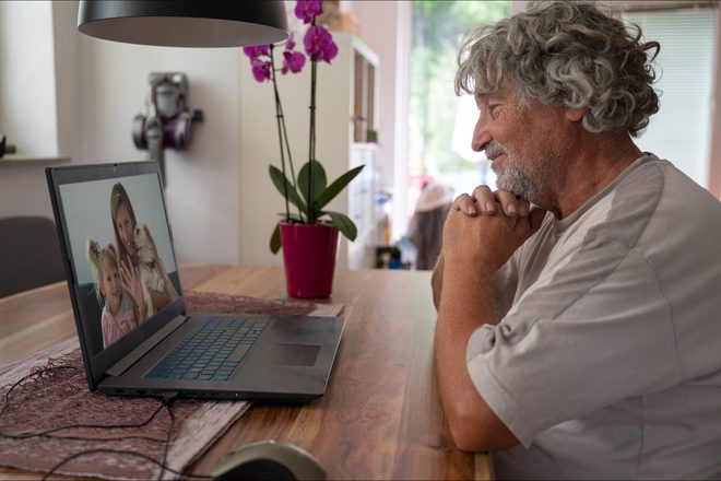 A smiling older man video chats on a laptop with a woman, a young girl and their dog.