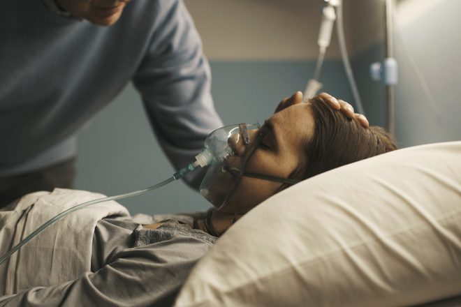 A man gently rests his hand on the head of a woman lying in a hospital bed with an oxygen mask.