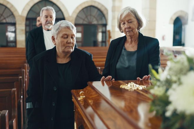 Two older women rest their hands on a casket in a quiet church.