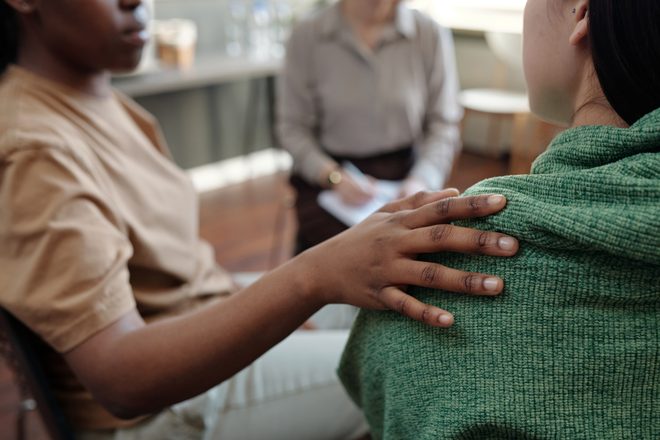 A woman rests a comforting hand on a friend's shoulder during a session with a counselor.
