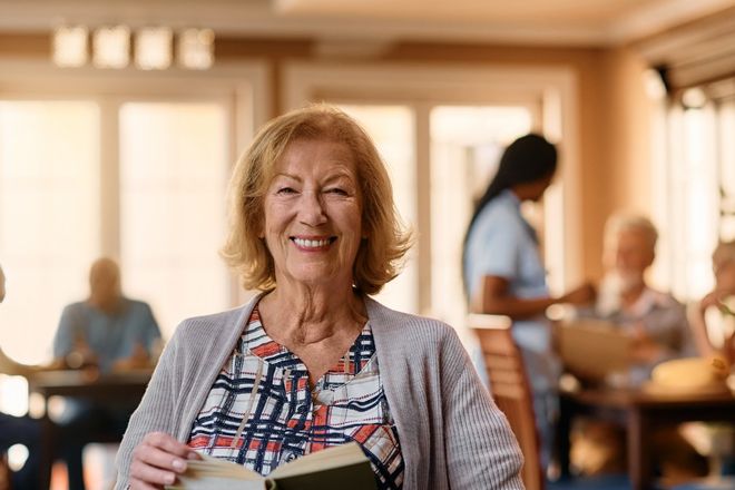A smiling older woman in a wheelchair holds an open book.