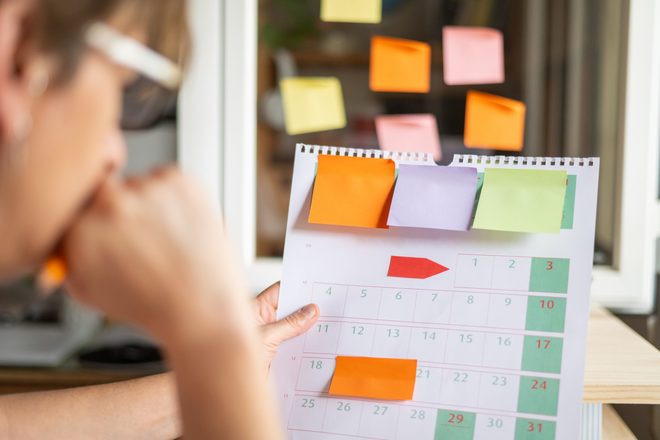 A woman studies a wall calendar marked with colorful sticky notes.