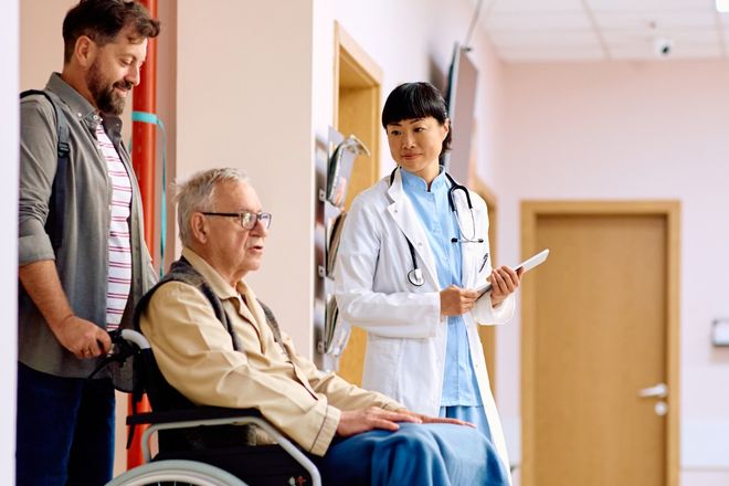 A man pushes his father in a wheelchair while a doctor talks with them in a clinic hallway.