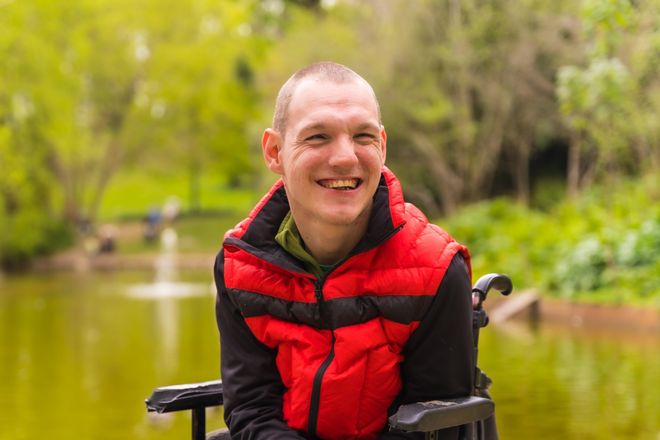 A smiling young man in a wheelchair by a pond in a park.